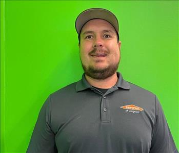 man in a grey SERVPRO shirt and hat standing in front of a green background. 